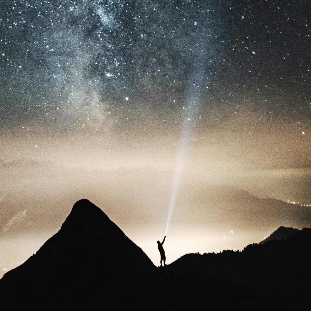 silhouette of person on top of mountain pointing flashlight on sky filled with stars at night time